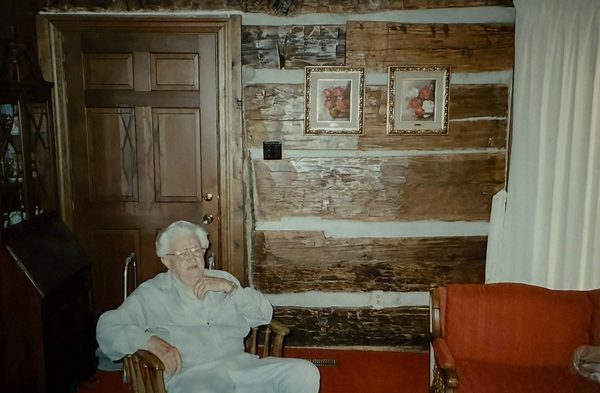 A man seated in a chair against a whitewashed log wall, beside a panelled door.