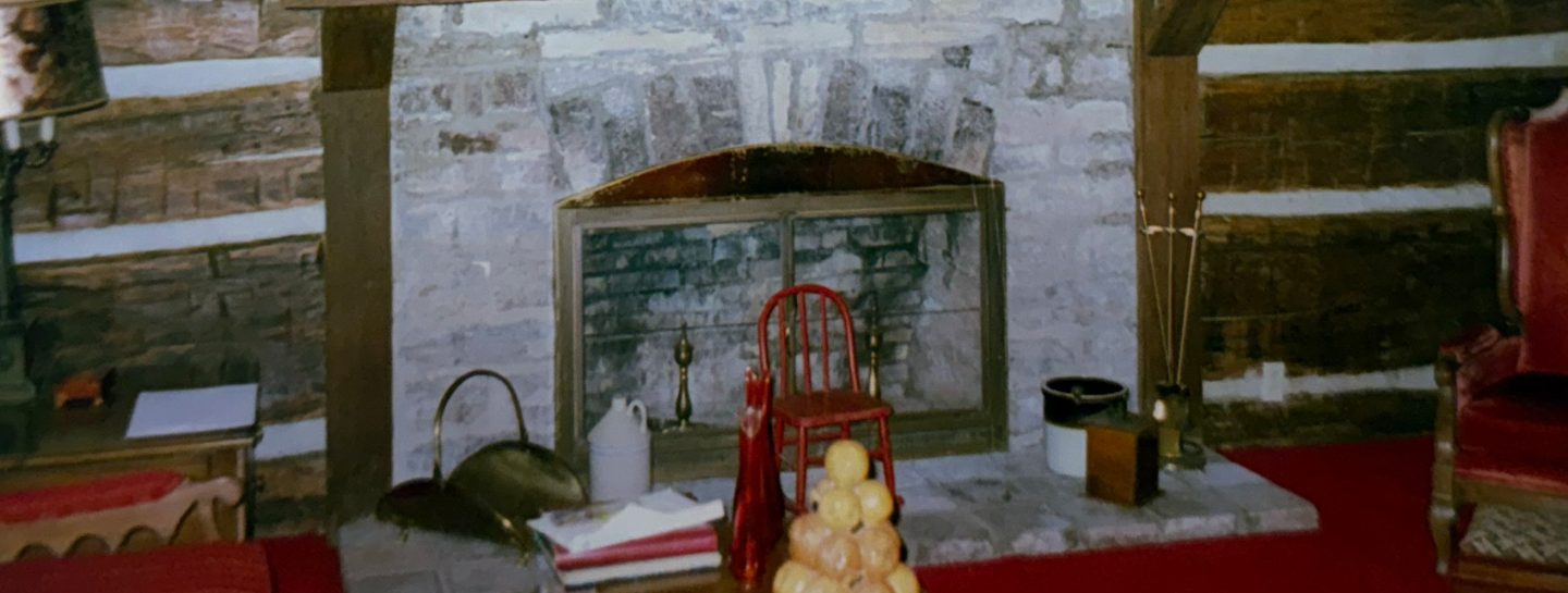 The stone fireplace and hearth inside the house, the log wall whitewashed between the courses.