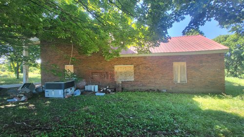 The side of the house under brick, its windows boarded, with a red metal roof.